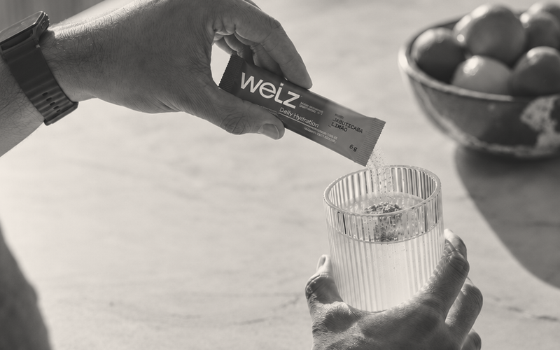 Mão despejando um sachê Welz Daily Hydration em um copo de água